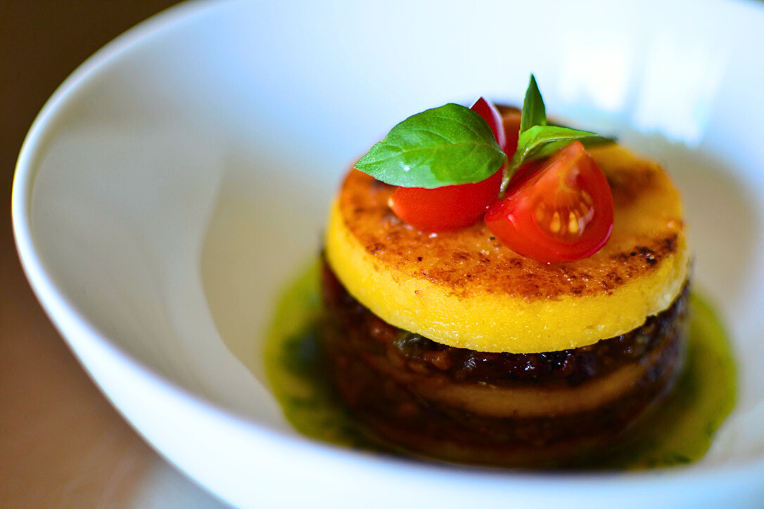 Bowl of three-tiered polenta cake appetizer with two tiers of tomato-bacon jam in a green herb oil